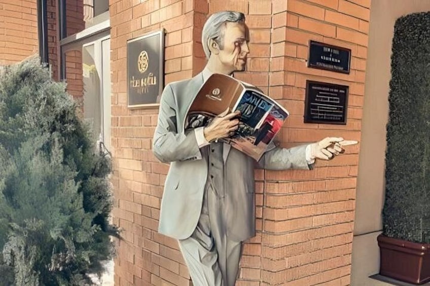 A statue of a man in a suit holding a book, standing next to a brick wall with plaques and a potted plant. 4