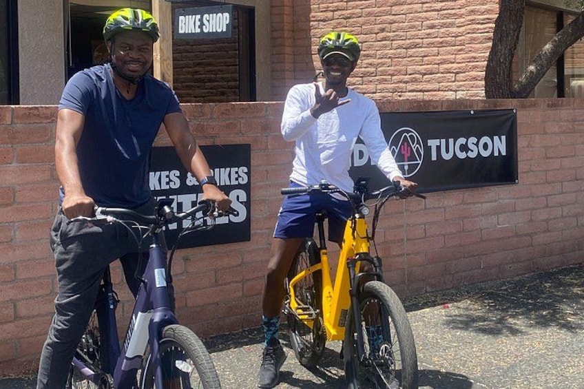 Two people on bicycles in front of a brick wall with signs for a bike shop and a location in Tucson. 1