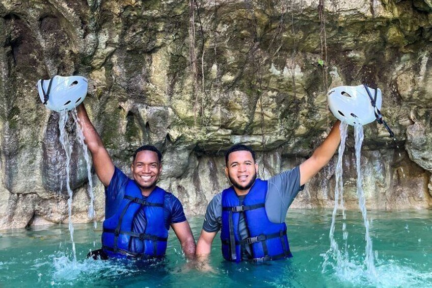 Two people in blue life vests holding white helmets above water in a rocky cave. 3