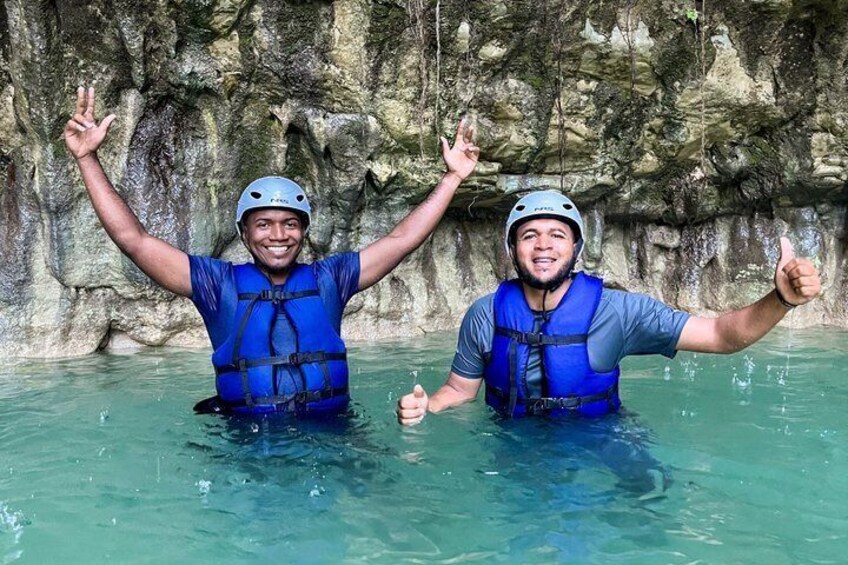 Two people in blue life jackets and helmets, standing in water with their arms raised, in front of a rocky cliff. 2