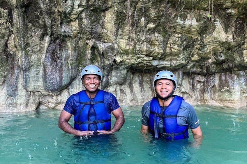 Two people in blue life vests and helmets standing in clear water with rocky cliffs in the background. 4