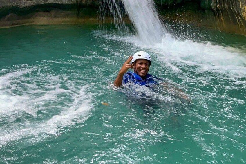 A person in a blue shirt and white helmet swimming in a turquoise body of water with a waterfall in the background. 5