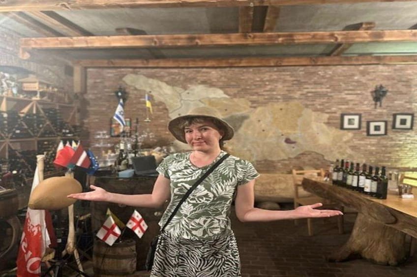 Una persona con vestido y sombrero a cuadros, parada en una bodega rústica con vigas de madera, botellas de vino y banderas. 5