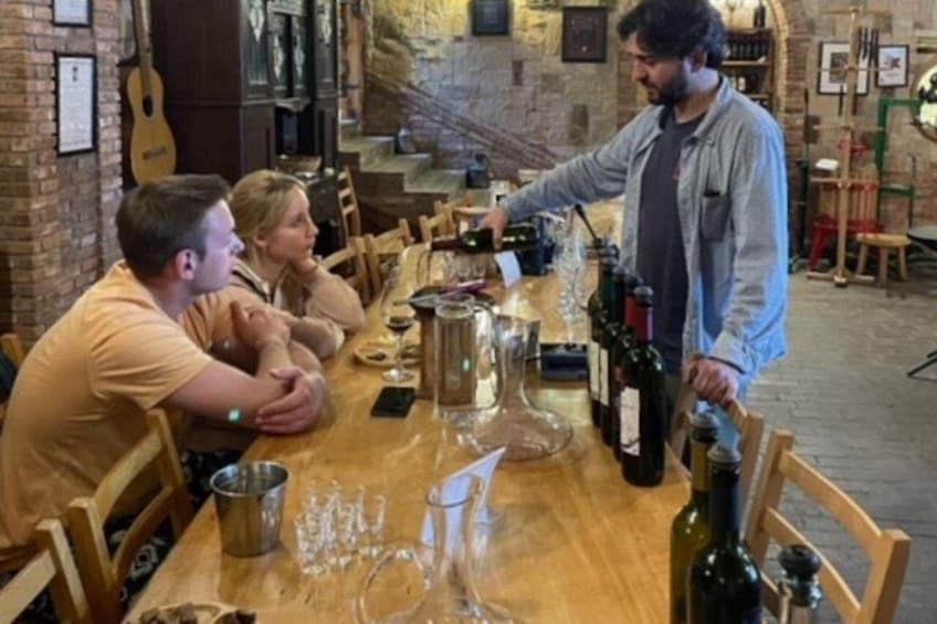 Una mesa de madera con botellas de vino y copas, una persona sirviendo vino, y un interior rústico con paredes de ladrillo y sillas de madera. 1