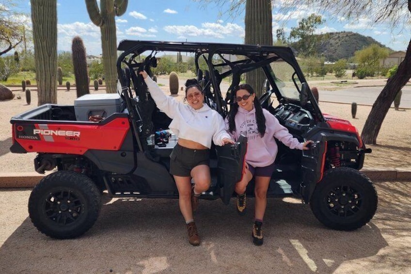 Two people sitting on a red and black Pioneer 4x4 vehicle in a desert with cacti and a clear sky. 1