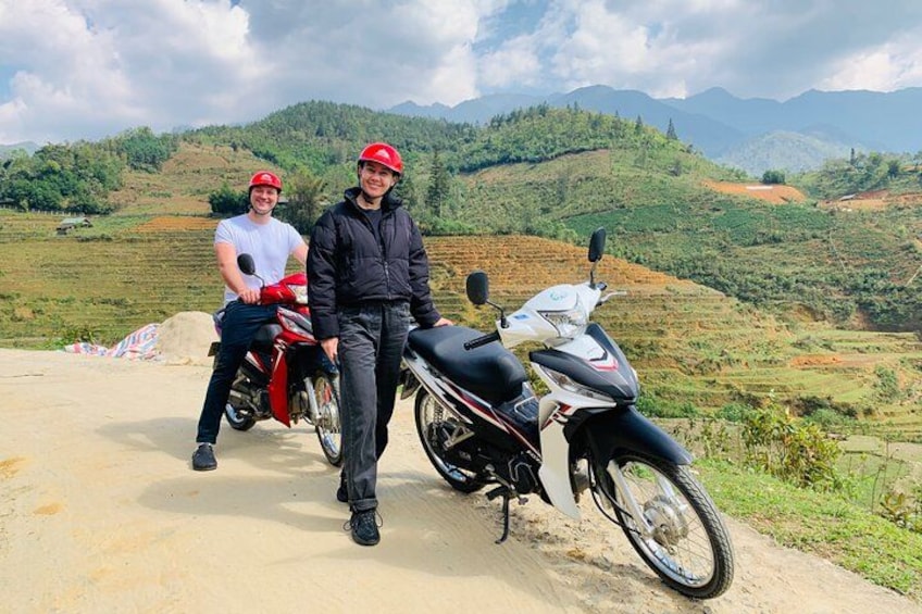 Two people standing next to motorcycles on a dirt road with a mountainous landscape in the background. 4