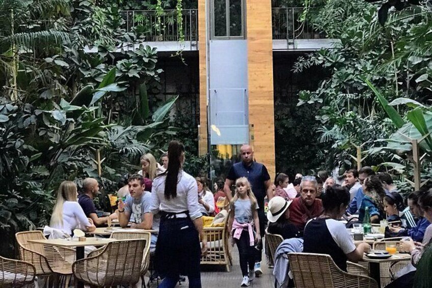 An outdoor dining area with wicker tables and chairs, surrounded by lush greenery and a modern building structure. 2