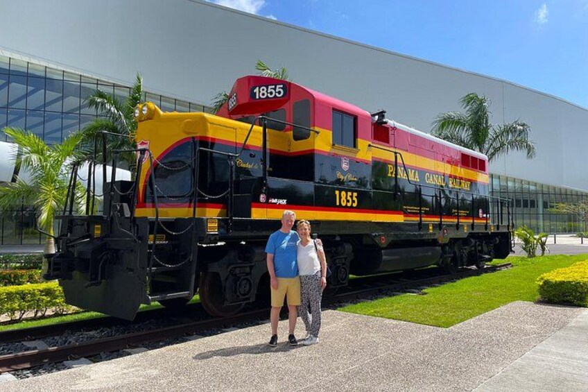 Een trein van de Panama Canal Railway met nummer 1855, een man en een vrouw staan voor een modern gebouw. 2
