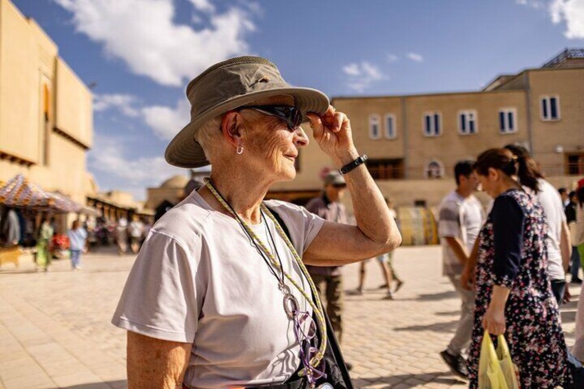 An elderly woman in a hat and sunglasses, standing in a public square with buildings and people in the background. 1