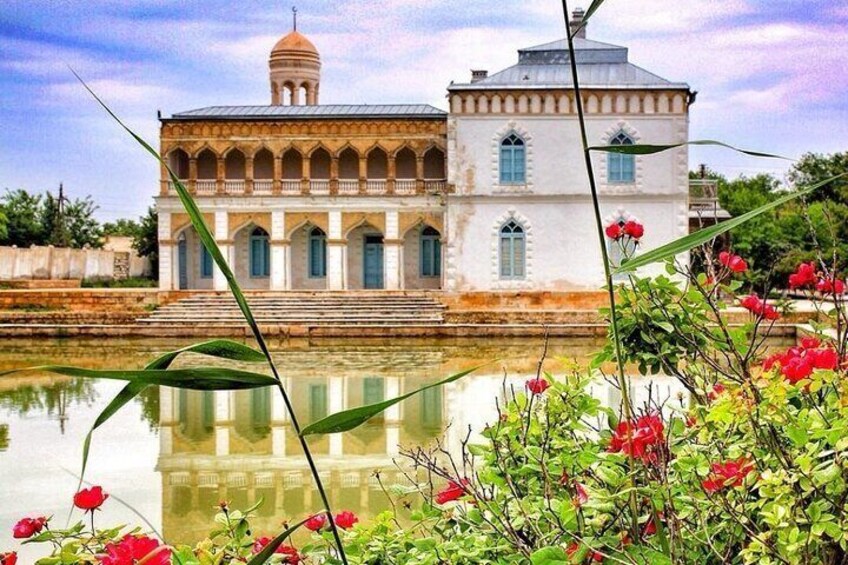 A historic building with a dome and arched windows, surrounded by a reflective water body and blooming red flowers. 1