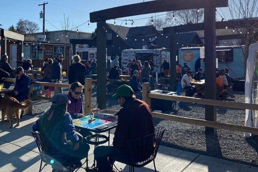 An outdoor market with wooden tables and chairs, people sitting and standing, and food stalls in the background. 1