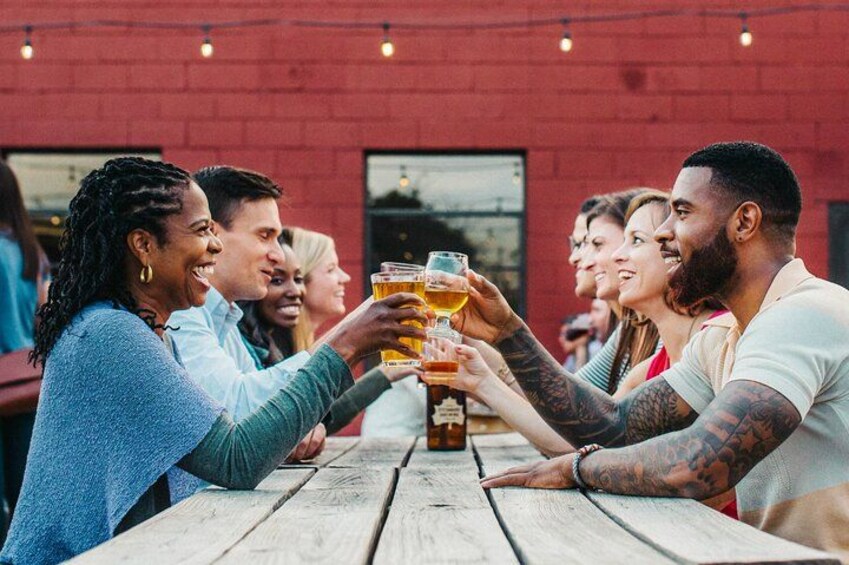 A group of people toasting with beer glasses at a wooden table outdoors. 2