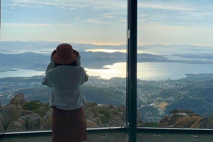 A person taking a photograph of a scenic view from a high vantage point overlooking a valley with a body of water and distant mountains. 3