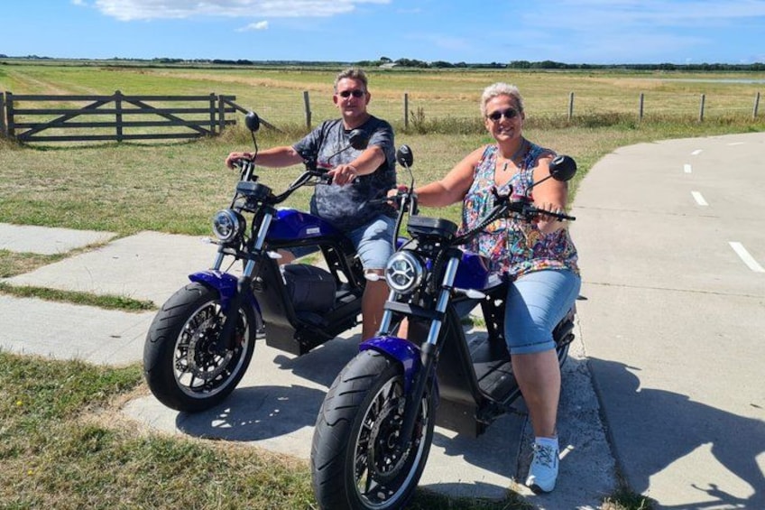 Two people on a blue motorcycle on a paved road with grass fields and a fence in the background. 1