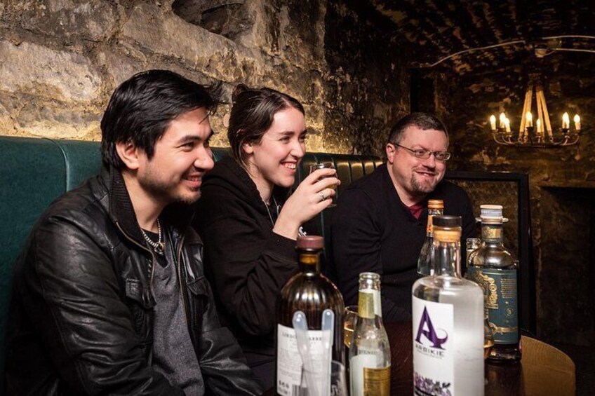 Tres personas sentadas a una mesa con botellas y copas, en una sala tenuemente iluminada con paredes de piedra y un candelabro. 4