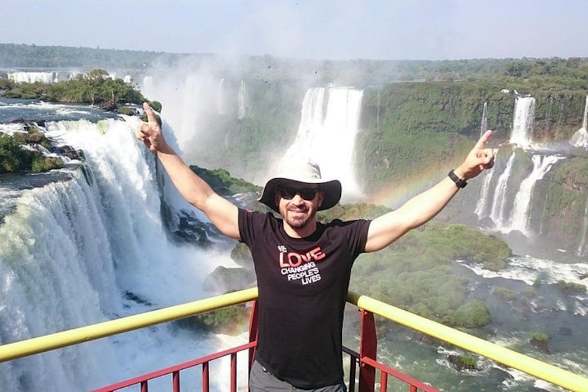 A person standing on a platform with arms raised, overlooking a large waterfall. 1