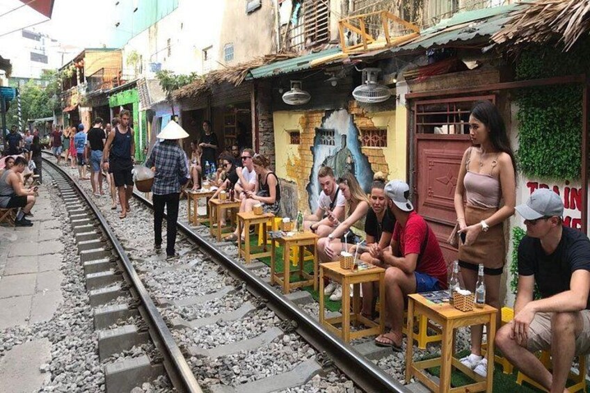 A train track with people sitting on benches, a woman in a conical hat, and a mural on a building. 2