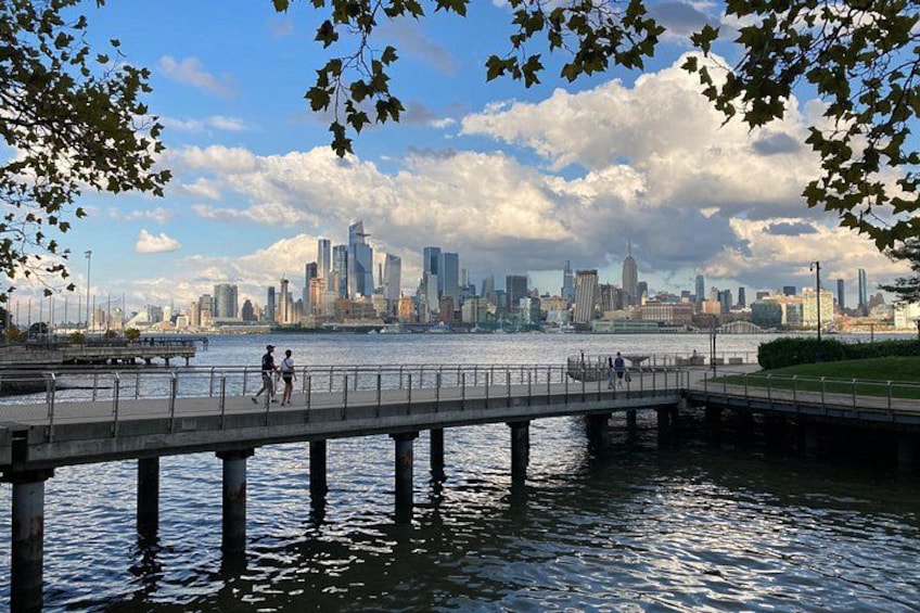 A waterfront with a pier, a city skyline in the background, and people walking. 1