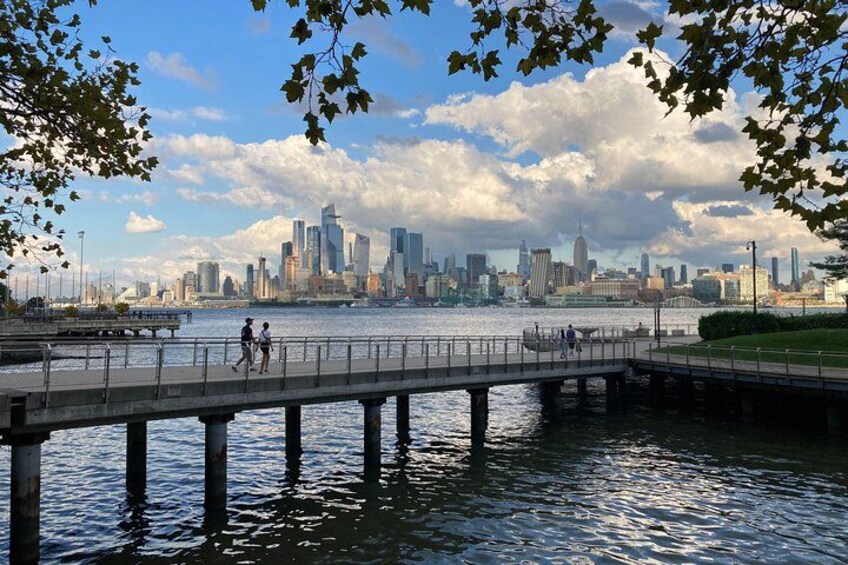 A river with a bridge, a city skyline in the background, and people walking. 1