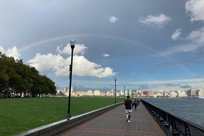A waterfront promenade with a rainbow, a city skyline, and a grassy area with trees. 4