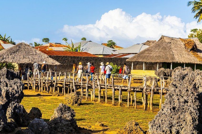 A rural village with thatched-roof huts, a wooden fence, and tourists. 4
