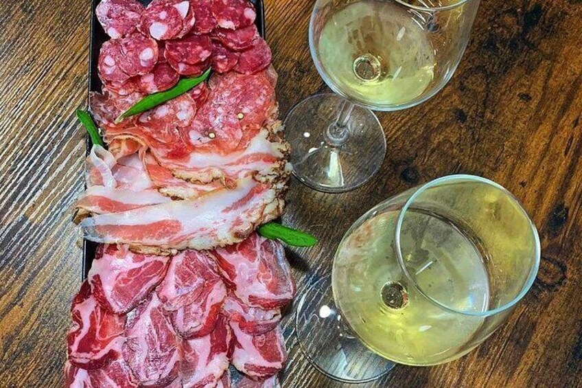 A charcuterie board with various meats and two glasses of white wine on a wooden table. 1