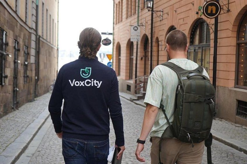 Two people walking down a cobblestone street, one wearing a dark blue shirt with a logo and the other carrying a backpack. 4