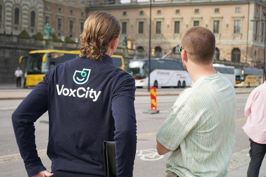 Two people wearing VoxCity branded jackets, a yellow bus, and a building with classical architecture in the background. 3