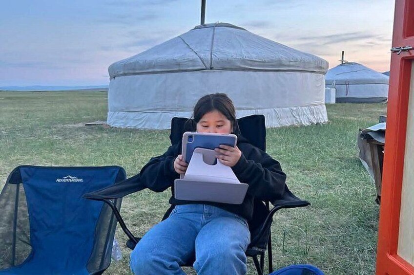 A person sitting in a chair with a laptop, a blue backpack, and a white tent in the background. 4