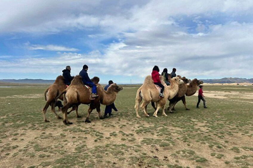 A group of people riding camels in a grassy field with a cloudy sky. 3