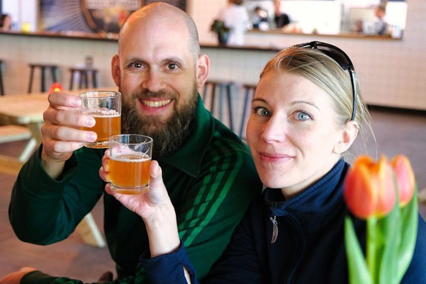Two people holding glasses of amber liquid, likely beer, in a casual indoor setting with a blurred background. 3