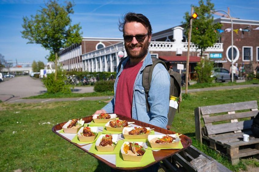A person holding a tray with food items outdoors, with buildings and a road in the background. 4