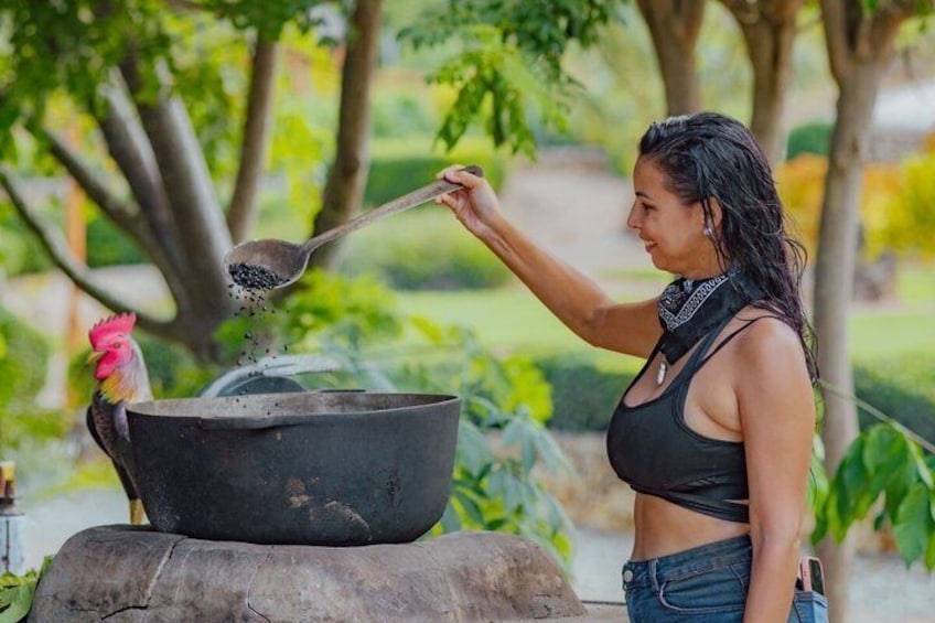 Una persona cucina all'aperto, con una pentola grande su un ceppo di legno, un gallo nelle vicinanze e vegetazione sullo sfondo. 1