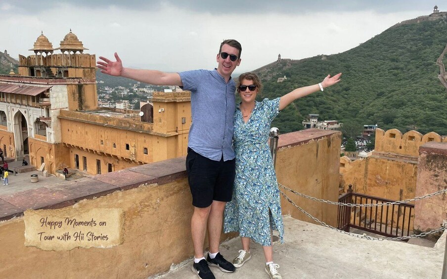 A couple standing on a balcony with arms outstretched, overlooking a historic fort and a hill in the background. 1