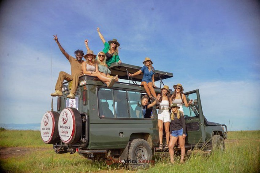 A group of people standing on and around a safari vehicle in a grassy field. 1