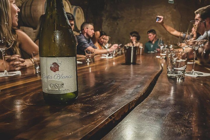 Une bouteille de vin Casta Blanca sur un comptoir en bois, avec des personnes assises autour. 1