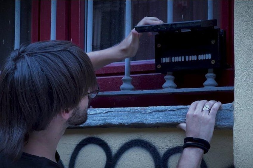 A person holding a camera with a black body and white buttons, standing on a balcony with a red railing and a window with white frames. 2