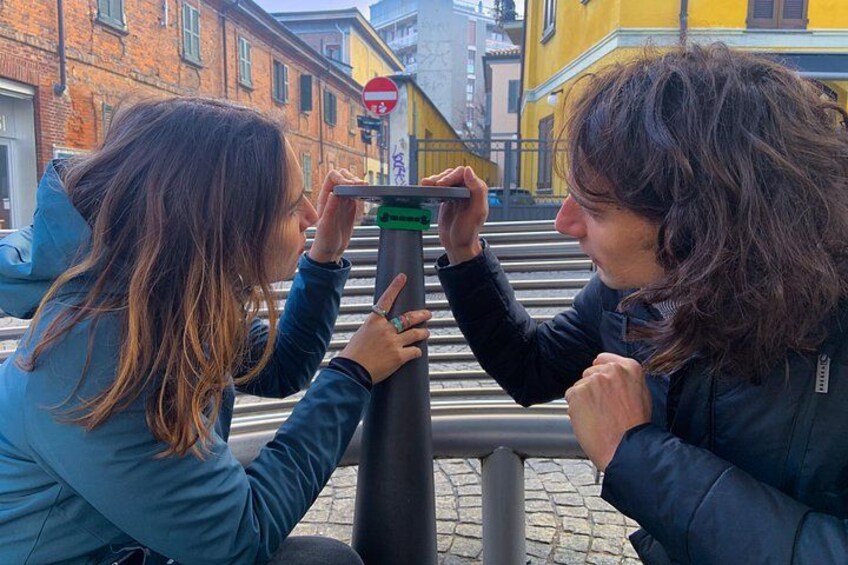 Two people using a telescopic device, possibly a binoculars, on a street with buildings and a no-entry sign in the background. 4