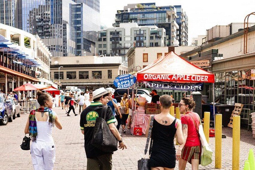 A busy street with people walking, a tent advertising organic apple cider, and buildings in the background. 5