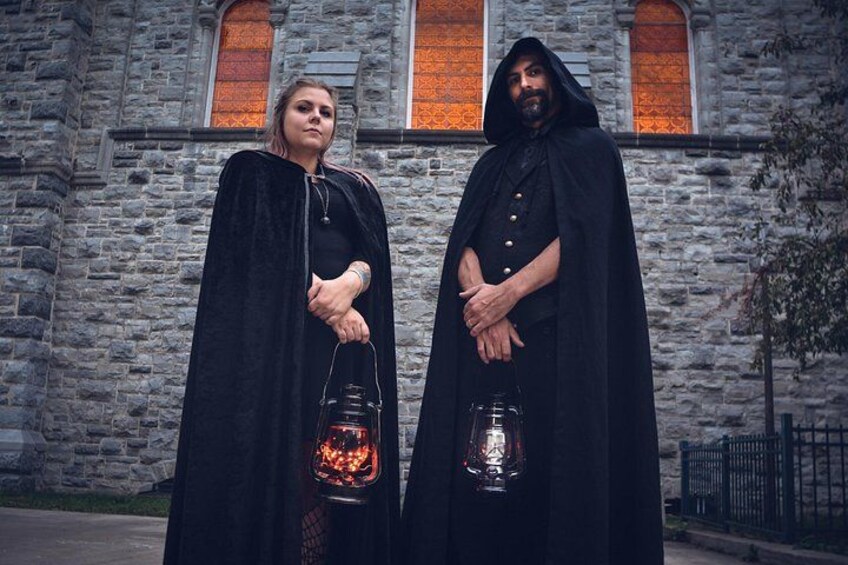 Two individuals in black cloaks standing in front of a stone building with arched windows. 2