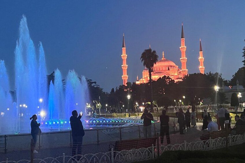 Une fontaine avec des jets d’eau, une mosquée aux minarets illuminés, et des gens qui se promènent. 3