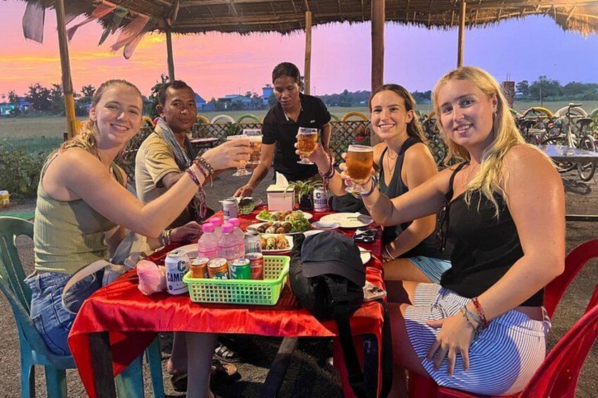 Eine Gruppe von Menschen sitzt an einem Tisch im Freien, mit Getränken und Snacks, unter einem Reetdach. 3