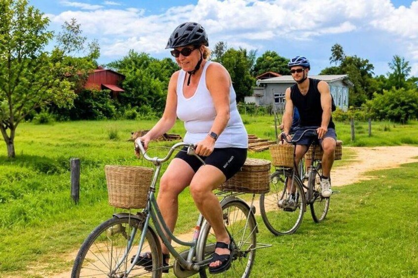 To personer cykler på en grusvej med kurve, omgivet af grønt og et hus i baggrunden. 3