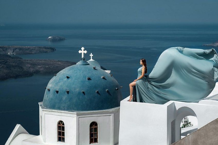 Une femme vêtue d’une longue robe fluide est assise sur une structure blanche surmontée d’un dôme bleu et d’une croix. Elle surplombe une étendue d’eau et un paysage rocheux. 5
