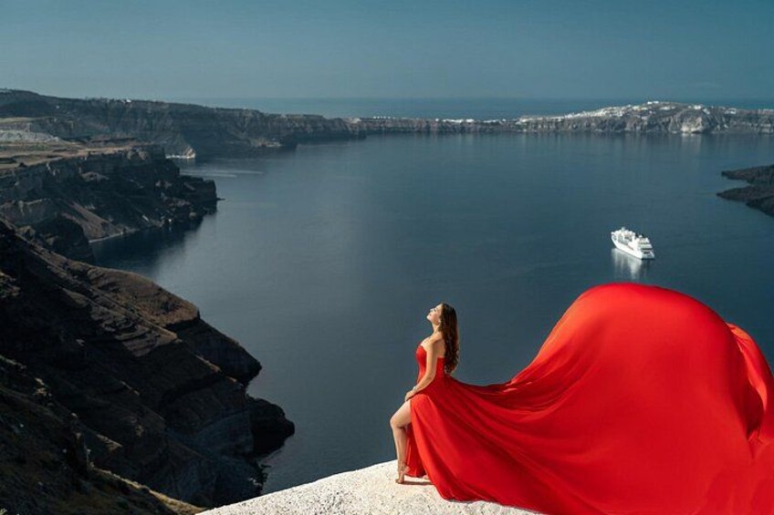 Une femme vêtue d’une robe rouge est assise sur une falaise, d’où elle contemple une mer calme, avec un bateau visible au loin. 2