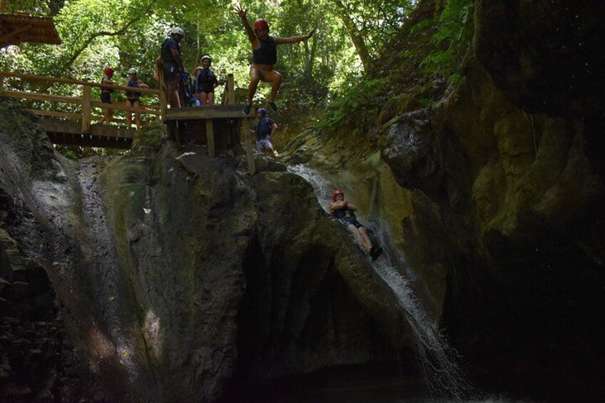 People on a wooden bridge in a forested area, with a person rappelling down a rocky cliff. 3