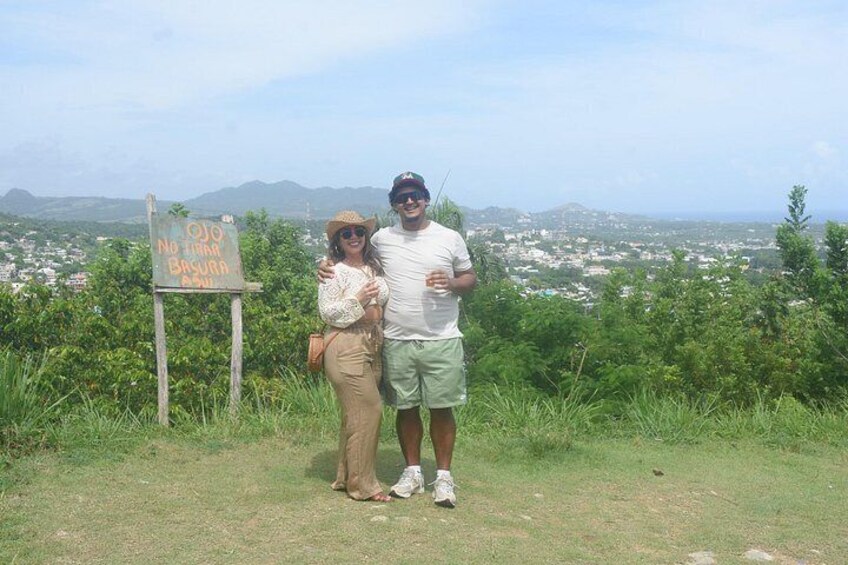 Two people standing on a grassy hill with a sign that reads 'OJO NO TENGO BASURA' and a cityscape in the background. 5