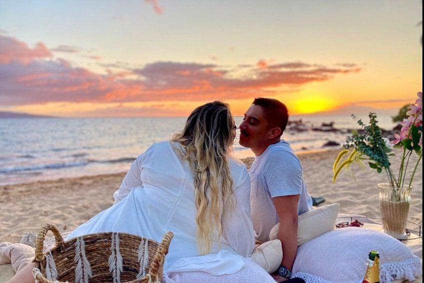A couple sitting on a beach at sunset with a basket and flowers nearby. 1