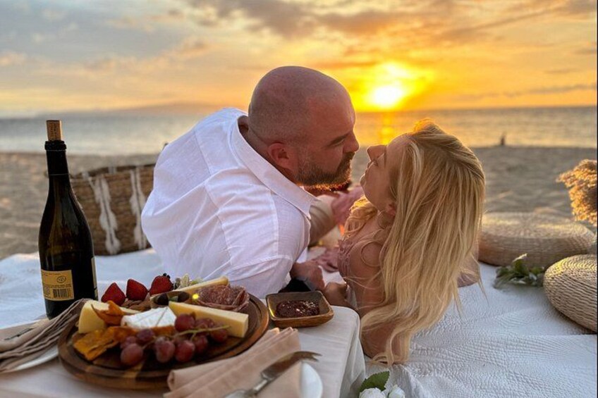 Una coppia su una spiaggia al tramonto, con un'area picnic che include formaggio, uva e una bottiglia di vino. 5