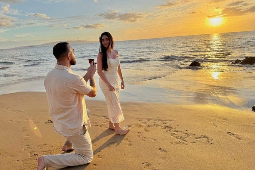 A person kneeling on a beach holding a ring, another person standing facing them, and the ocean in the background. 2